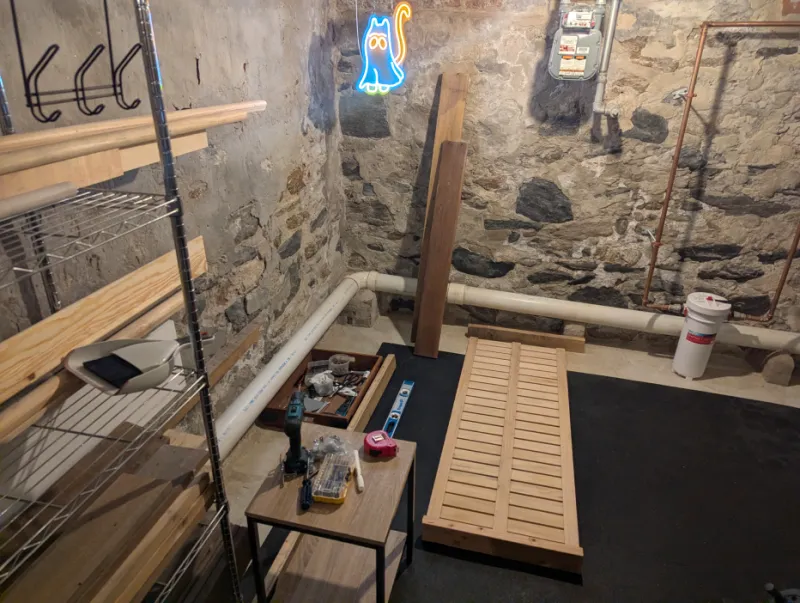 Daybed slats and lumber laid out on a basement floor mat beside a side table covered in tools